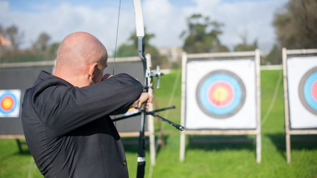 A bald man in a suit practices archery on a sunny day in Portugal, aiming at targets.